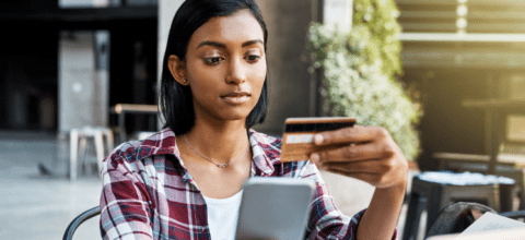 Young woman looks at a credit card while holding a phone on her right hand.
