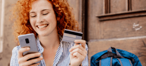 A woman smiles looking at a mobile phone while holding a credit card in her left hand.