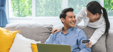 A man sitting on a couch with a laptop smiles while looking at a woman leaning in from behind the couch while holding a credit card.