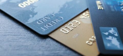 A close-up image of three stacked credit cards on a gray surface. The cards are slightly fanned out, revealing parts of each card, including visible digits, embossed text, and partial world map designs.