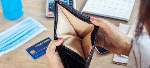 Person is holding open their empty wallet. Behind the wallet, on the table, lay a credit card and blue face mask.