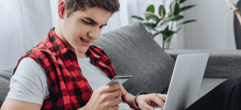 Young man smiling and sitting on couch with a laptop and holding a credit card in his hand.