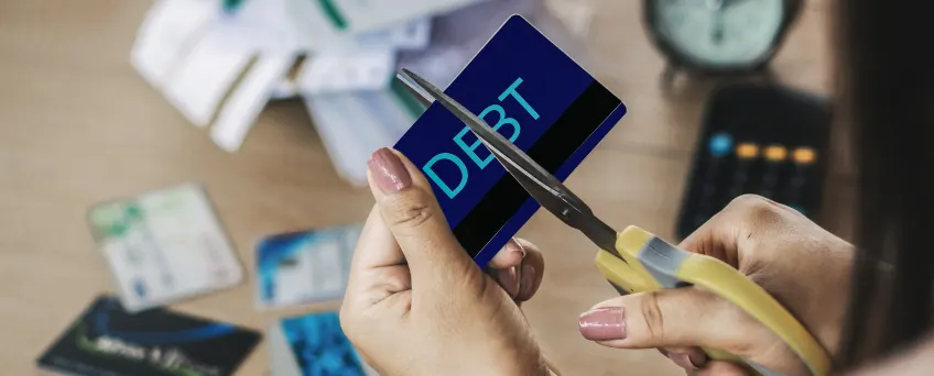 A person is using scissors to cut a credit card labeled DEBT. In the background, various cards and papers are scattered on a table. A blurred clock and calculator are also visible.