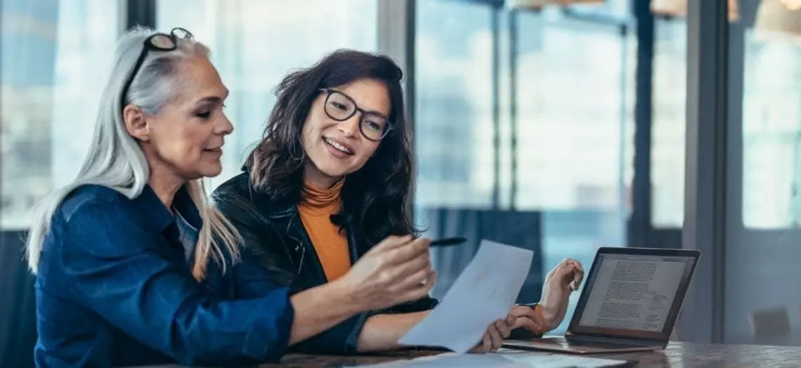 Two women are sitting at a table in an office setting. One is holding and pointing at a document, while the other smiles and listens. A laptop sits open with text visible on the screen. Both appear engaged in discussion.
