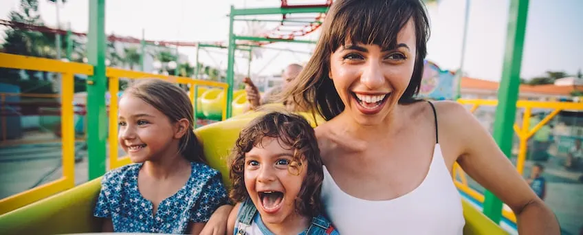A woman and two children enjoy a roller coaster ride. They are seated in a yellow coaster car, laughing and smiling with excitement. The background shows the blurred tracks and colorful structures of the amusement park.