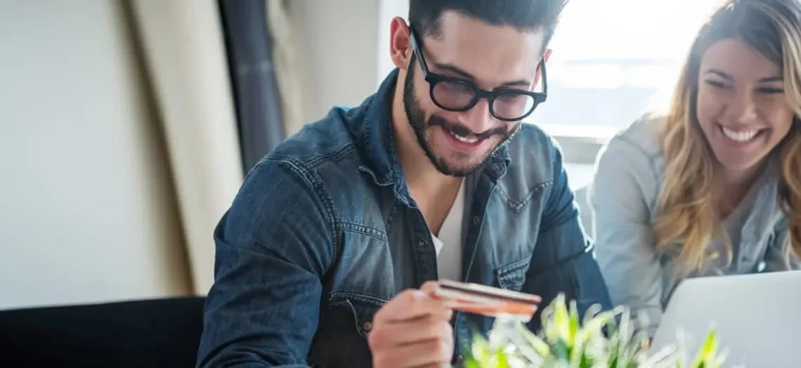 A laughing couple looks at their credit card while in front of a laptop.