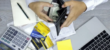 A person is organizing dollar bills in a black wallet. The scene includes a notebook, credit cards, a laptop, a calculator, and sticky notes on a wooden table, suggesting a financial planning or budgeting context.